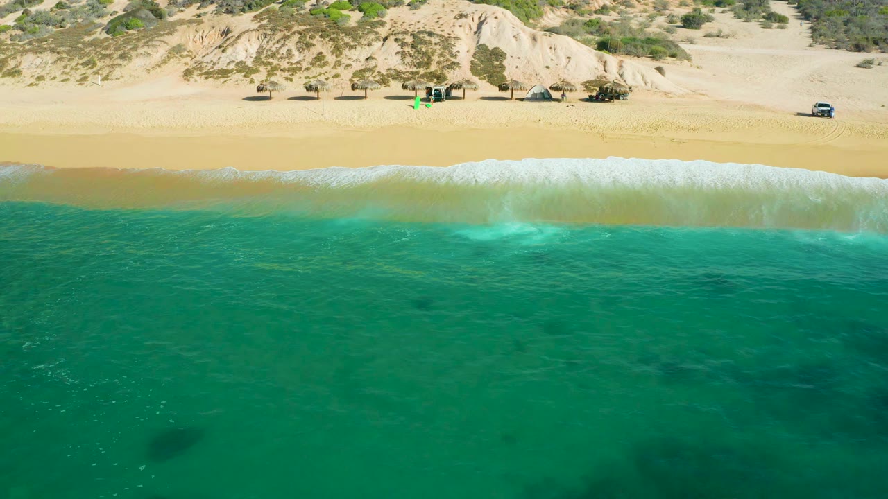 Aerial frame at Shipwrecks Beach, Baja California Sur, with the surf band slightly farther up the sand, clear green-blue water in front, dune slopes behind, and no visible people.