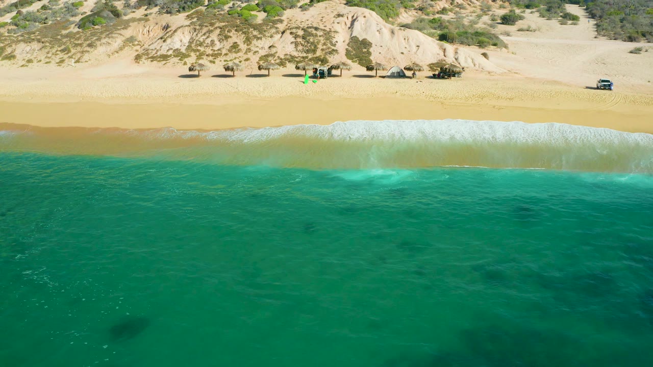 Aerial frame at Shipwrecks Beach, Baja California Sur, showing shallow turquoise water with darker underwater patches, a thin foam line, open beach sand, dune vegetation, and no visible people.
