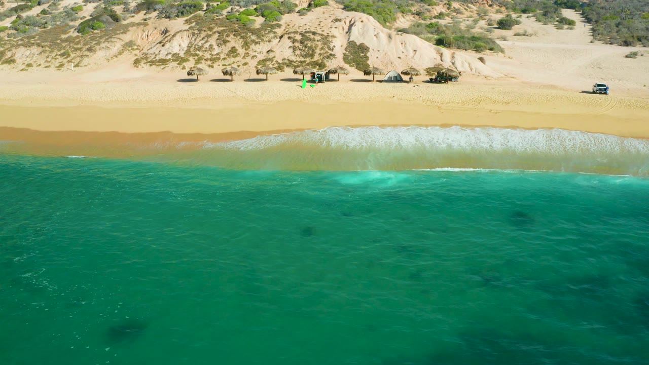 Aerial frame at Shipwrecks Beach, Baja California Sur, with green water across the foreground, white surf breaking onto smooth sand, dune ridges in back, small vehicles near the beach, and no visible people.