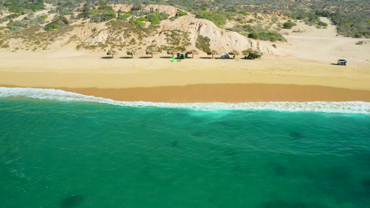 Opening aerial frame over Shipwrecks Beach in Baja California Sur, with turquoise water in front, a thin white surf line, golden sand, low dunes, scattered beach structures, vehicles, and no visible people.