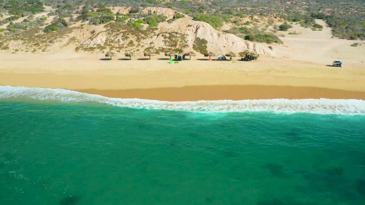 Shipwrecks Beach aerial frame with broad turquoise water below, foamy surf curling onto a wide sandy strip, pale eroded dunes behind it, scattered shade structures, vehicles, and no visible people.