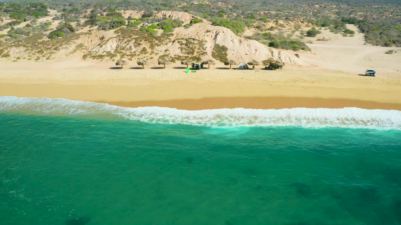Mid-clip aerial frame over Shipwrecks Beach with teal water taking most of the foreground, a brighter breaking wave at the shore, golden beach, dune ridges, sparse scrub, small vehicles, and no visible people.
