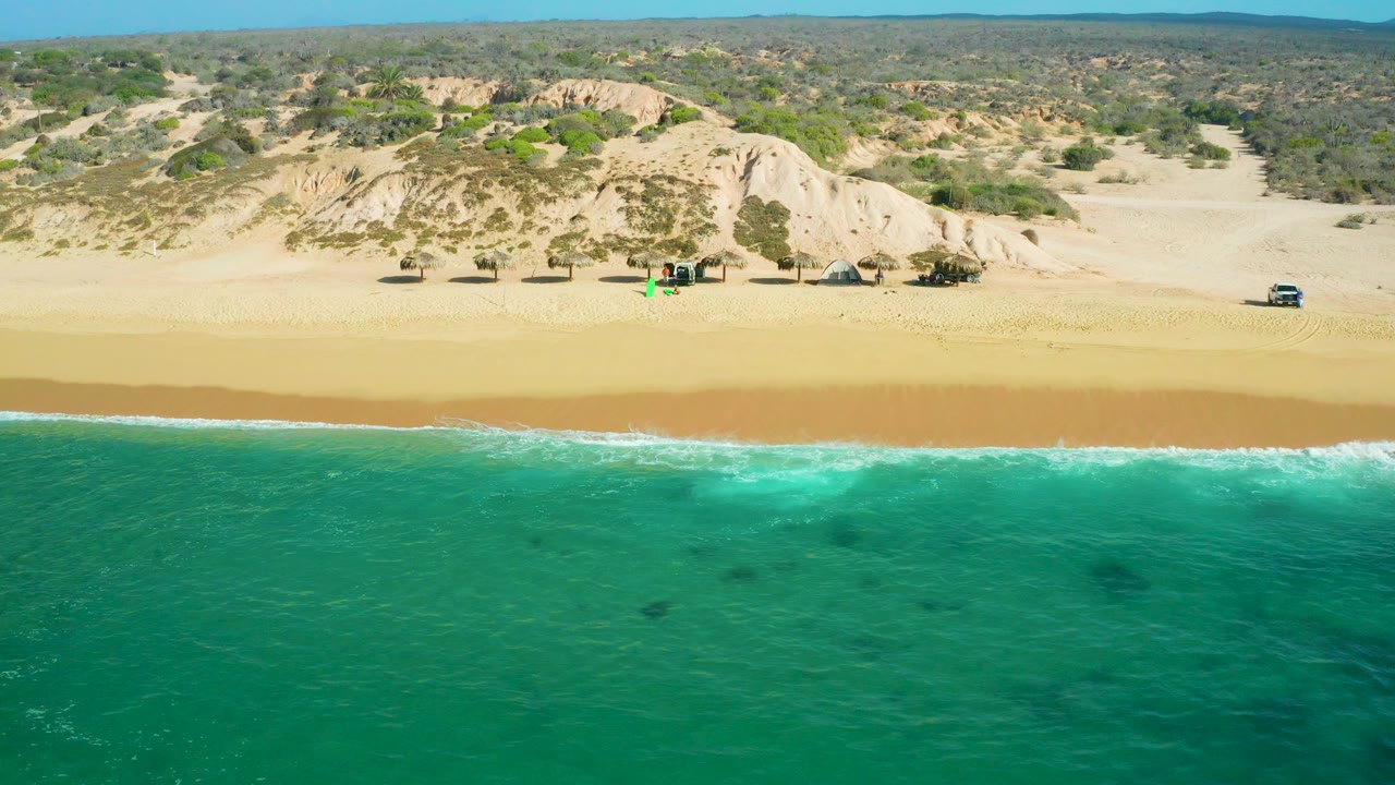 Aerial view of turquoise water, surf, golden beach, dunes, and scrubland.