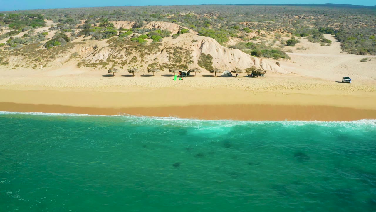 Aerial view at Shipwrecks Beach with darker patches in the turquoise water, a broad strip of sand, dune slopes behind it, scattered scrubland, and no visible people.