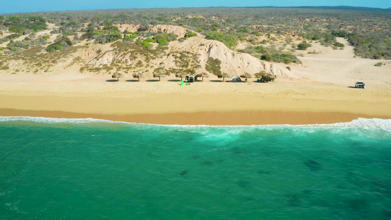 Frame from Shipwrecks Beach showing a longer white wave breaking across the lower shoreline, golden sand through the center, pale dune banks, scrubland, distant hills, and no visible people.
