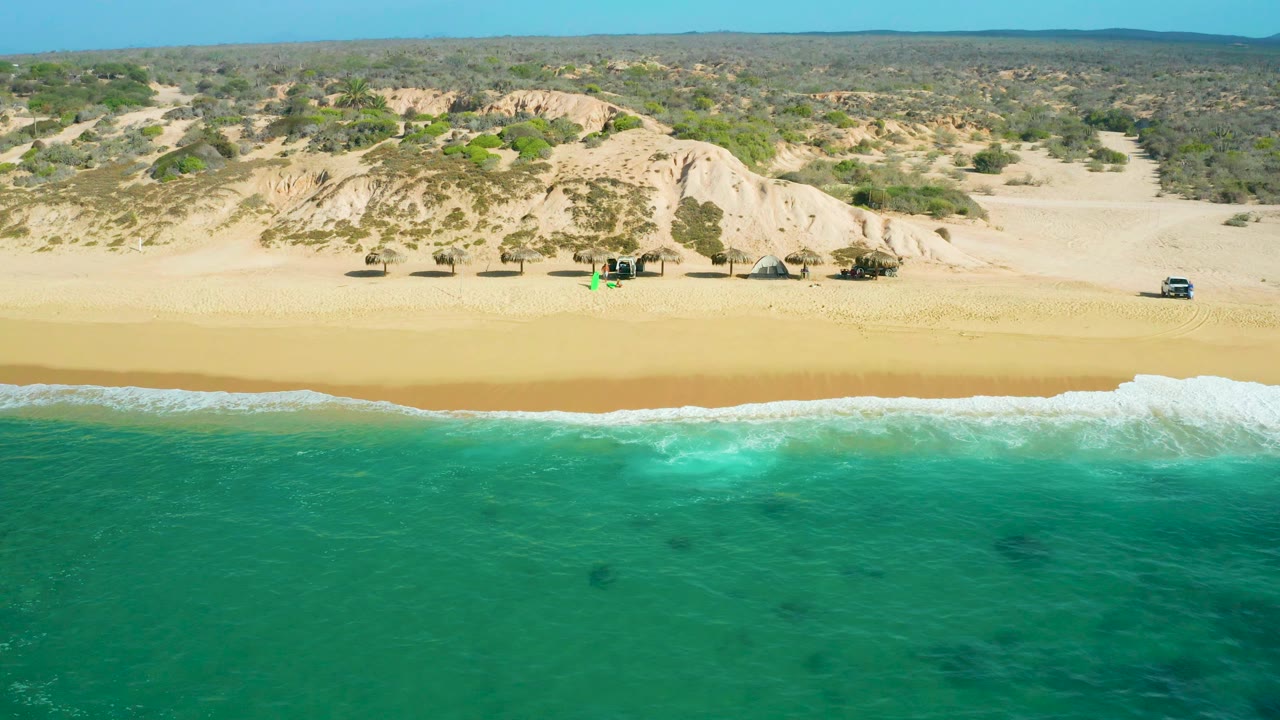 Aerial frame at Shipwrecks Beach with foamy surf crossing the foreground, an open sandy beach, a light-colored dune ridge, scattered coastal plants, distant low hills, and no visible people.