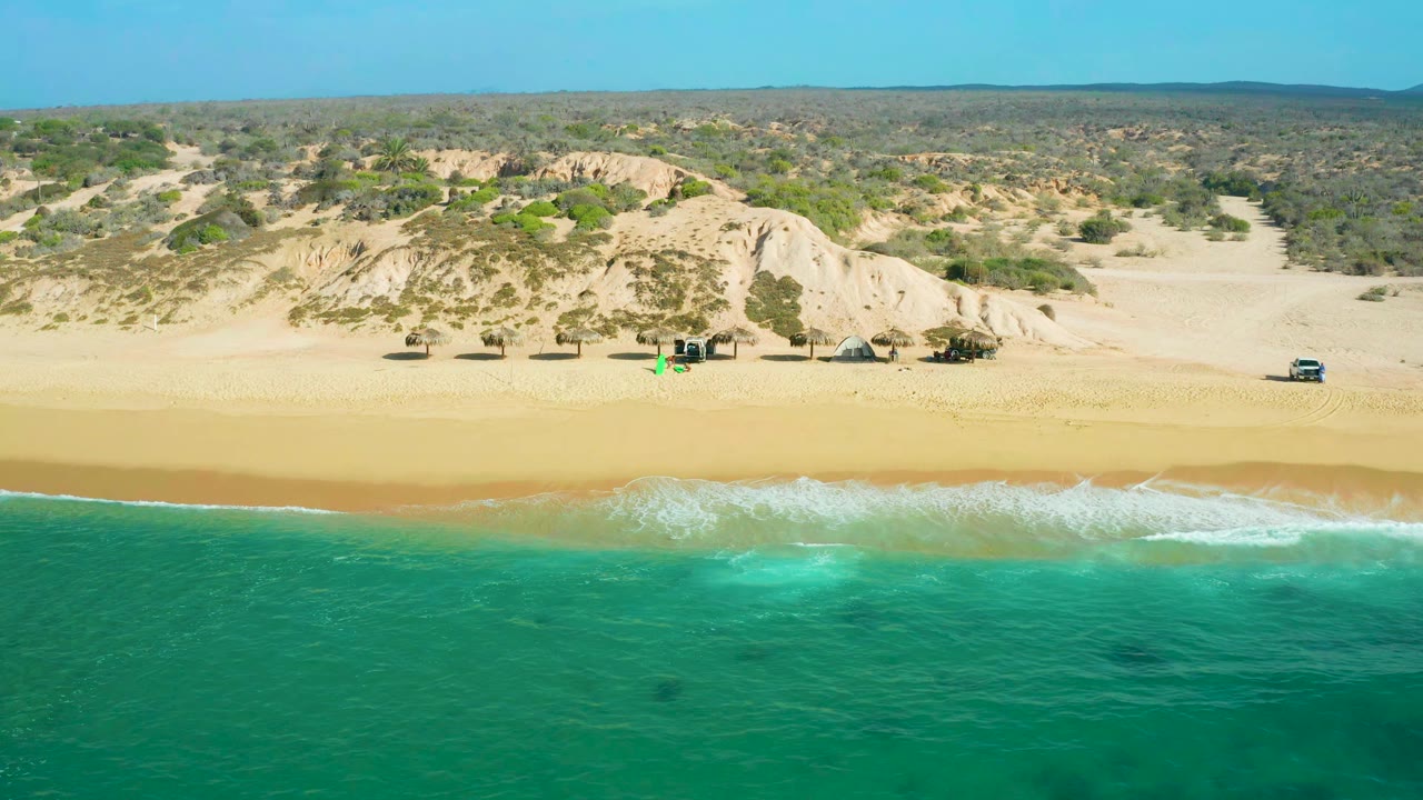 A second aerial frame shows white foam spreading across the shoreline at Shipwrecks Beach, with tan sand, dune slopes, low vegetation, and no visible people.