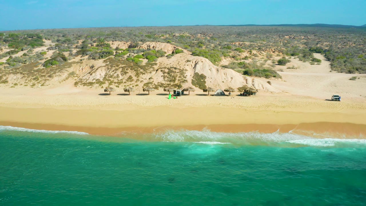 The frame holds a broad aerial composition of Shipwrecks Beach, with darker green water in front, a fresh surf band on the sand, pale dunes, scrubland, and no visible people.