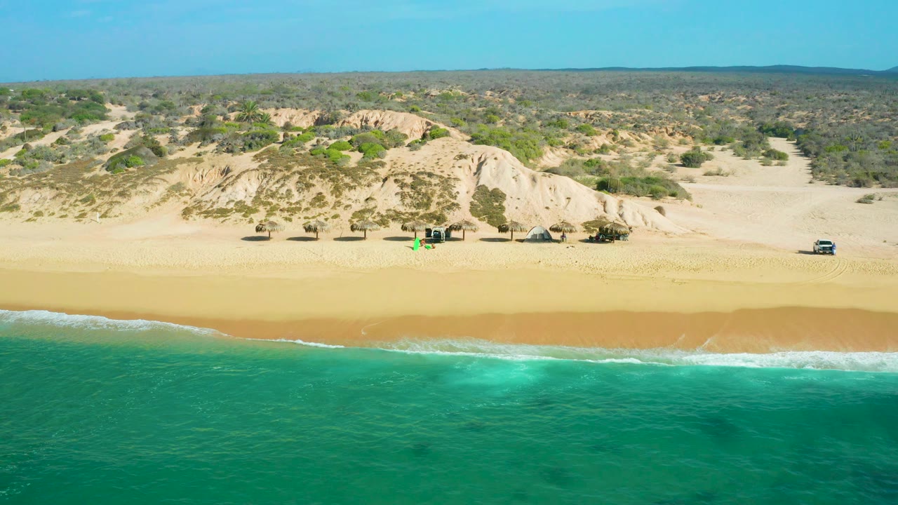 At 0:03, Shipwrecks Beach appears from above with foamy surf sliding along the lower edge of the sand, dunes and brush beyond, and no visible people.