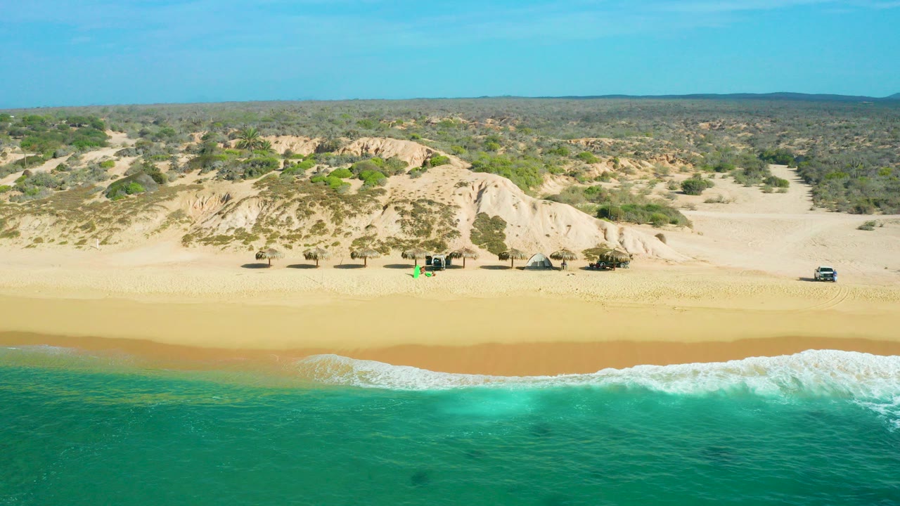 Opening aerial frame over Shipwrecks Beach in Baja California Sur, Mexico, with turquoise water in the foreground, tan sand, pale dunes, sparse beach vehicles, desert scrub, and no visible people.