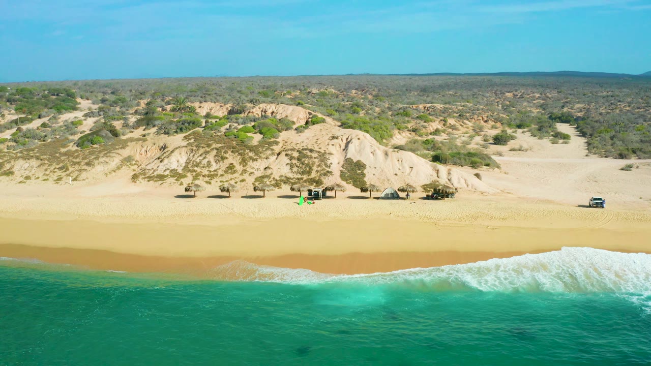 High-angle drone frame at Shipwrecks Beach showing a larger white surf band entering from the right, open sand, pale dune slopes, scrubland behind the beach, parked vehicles, and no visible people.