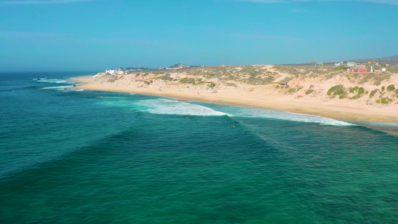 Opening aerial frame at Shipwrecks Beach, Baja California Sur, showing deep blue foreground water, a pale surf line, sandy shoreline, low dunes, scattered buildings, and no visible people.