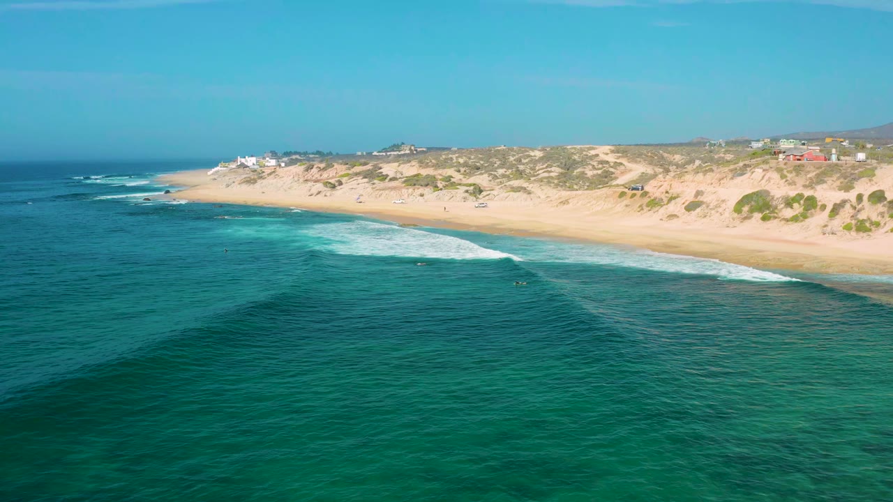 Aerial view over blue-green water at Shipwrecks Beach, Baja California Sur, with a thin white wave breaking along the sand and no visible people in the frame.