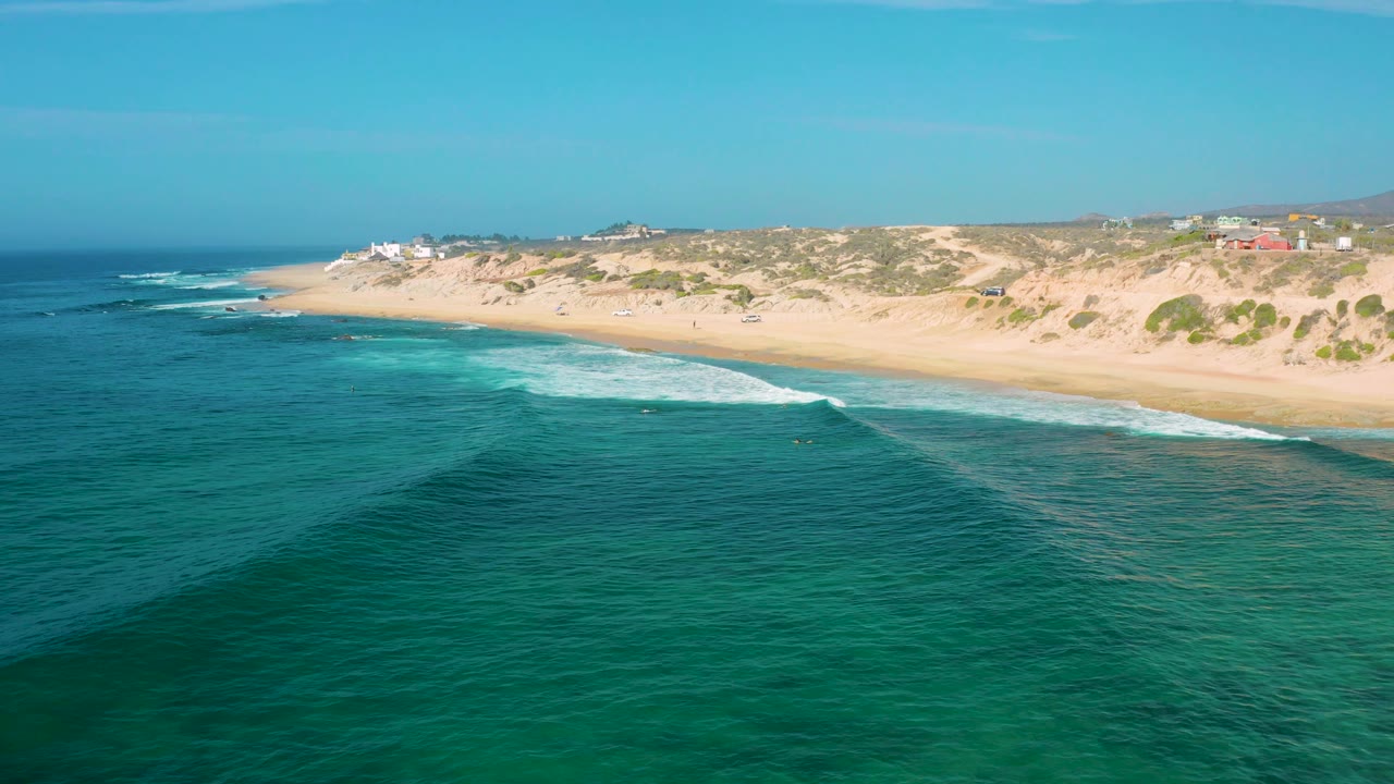 Shipwrecks Beach aerial frame with turquoise shallows near the surf, deeper blue water in front, a long golden beach behind, and no visible people.