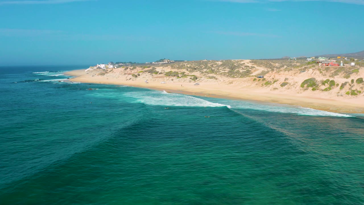 Aerial frame from Shipwrecks Beach, Baja California Sur, with calm blue water in the foreground, a white wave strip crossing the midground, sandy dunes behind, and no visible people.