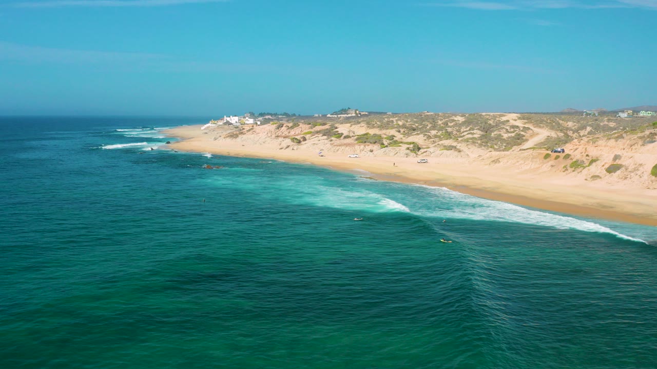 Aerial view over Shipwrecks Beach in Baja California Sur, with turquoise water in the foreground, a diagonal white surf line, dunes beyond the sand, and no visible people.