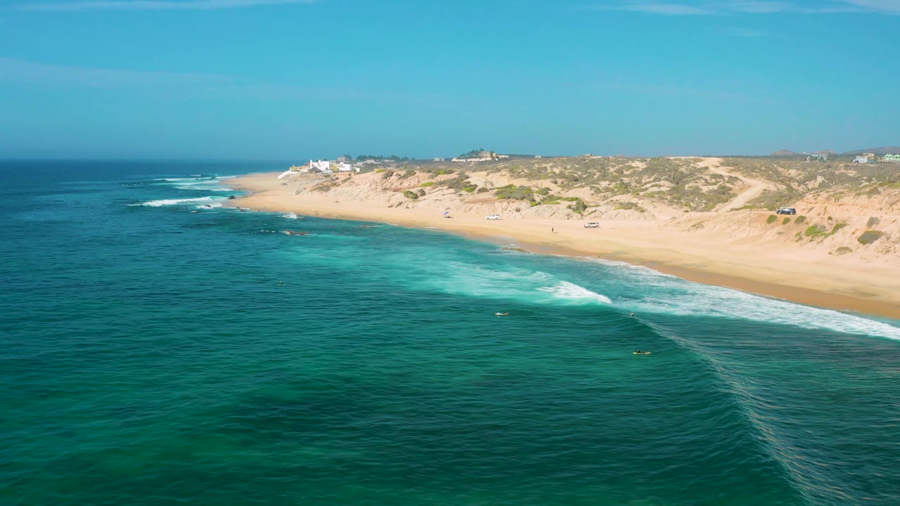 Drone view over Shipwrecks Beach in Baja California Sur, showing darker offshore water, pale turquoise shallows, white surf bands, sandy dunes, and no visible people.