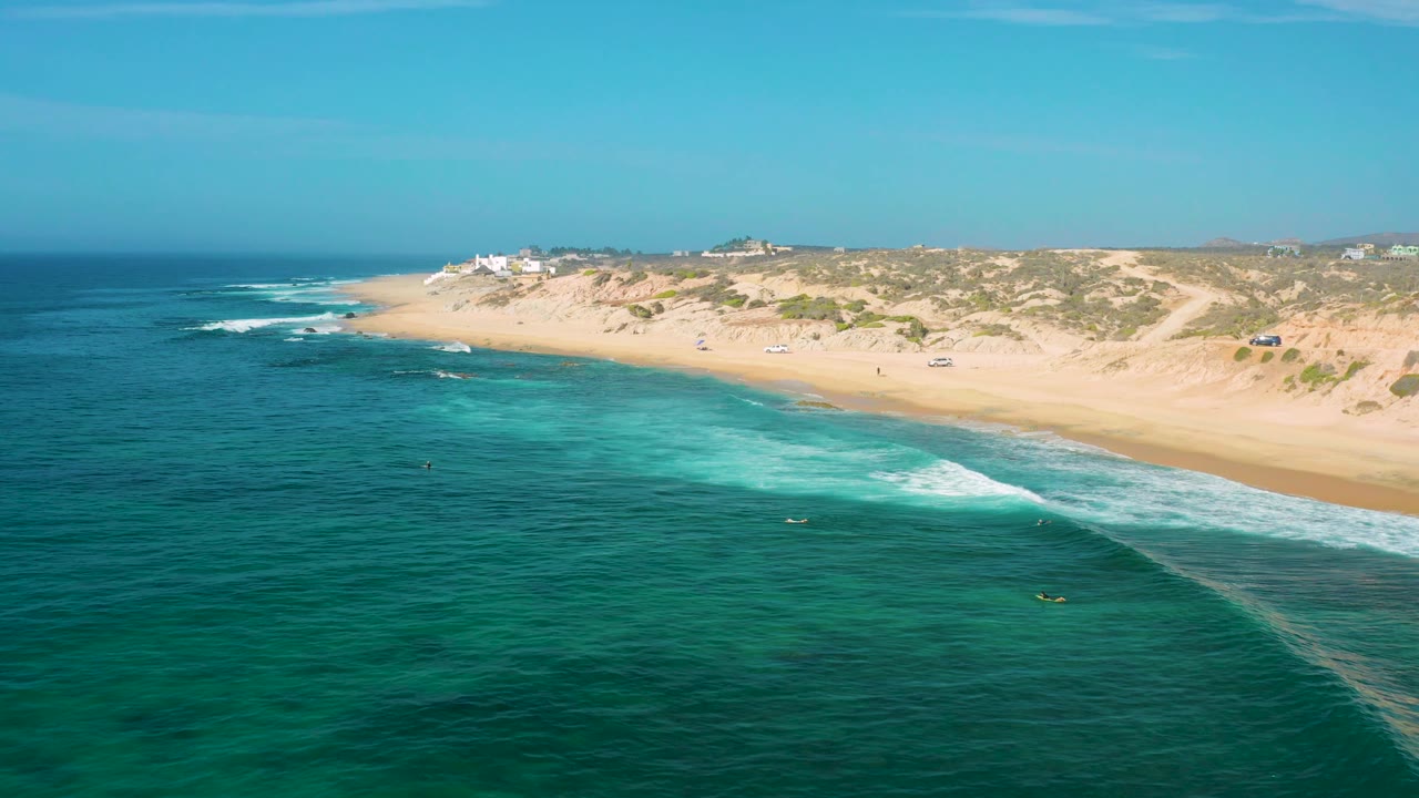 Aerial view at Shipwrecks Beach, Baja California Sur, with a foamy wave breaking close to the sand, low dunes behind the beach, distant buildings, and no visible people.