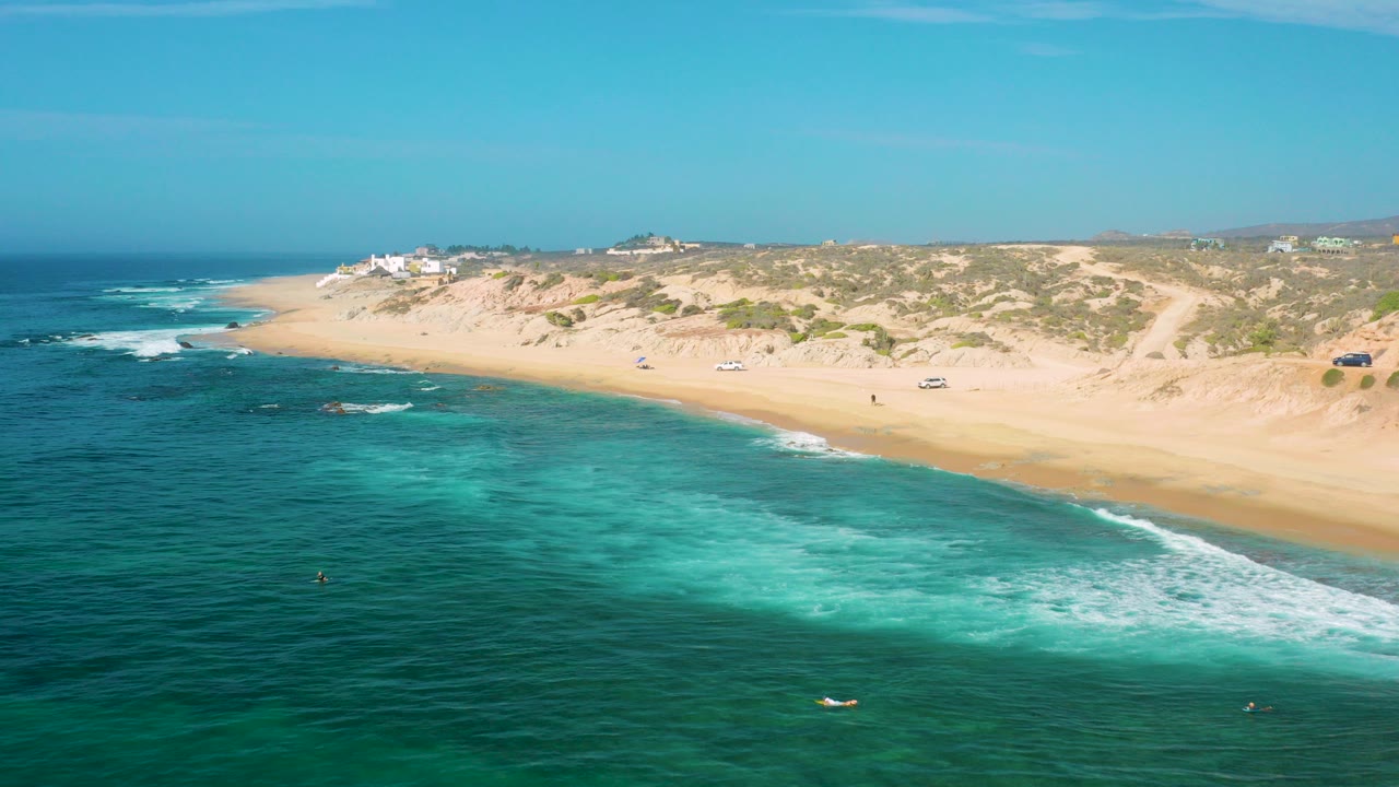 Aerial view over Shipwrecks Beach in Baja California Sur, with turquoise water in the foreground, a broad sandy shoreline, dune scrub, small buildings, and tiny people visible on the beach.