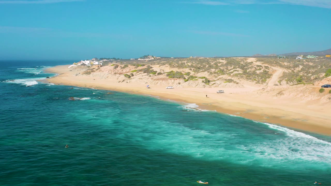 Aerial view along Shipwrecks Beach where a broad patch of pale turquoise water sits beside darker ocean, with white foam, sandy beach, dunes, and small structures on the horizon.