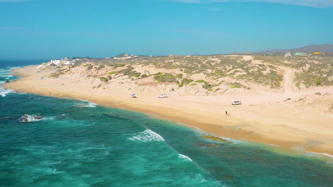 Aerial view over Shipwrecks Beach in Baja California Sur, with turquoise water in the foreground, a sandy shoreline, dune vegetation, parked vehicles, and small beachgoers in the distance.