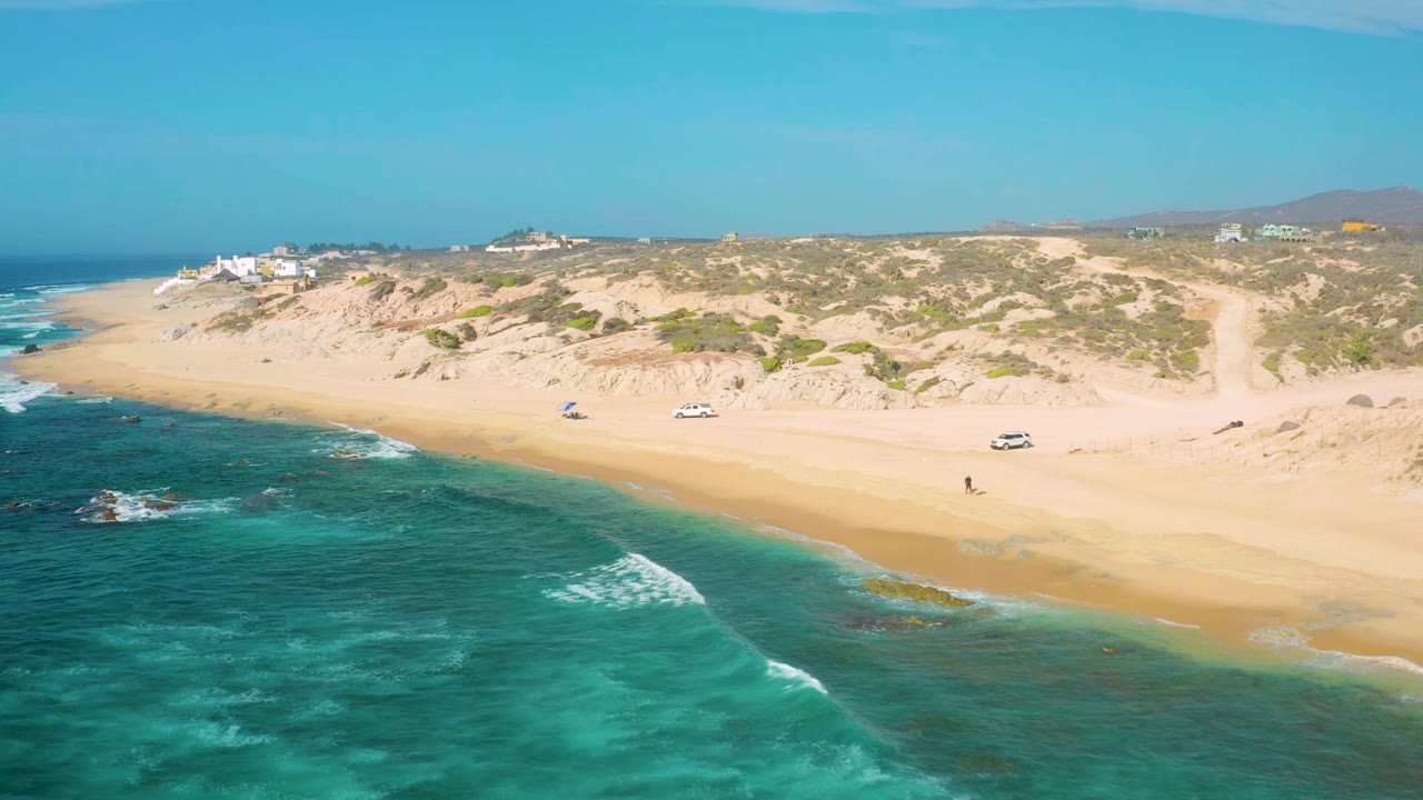 Aerial frame at Shipwrecks Beach showing shallow green-blue water, a broad strip of sand, two visible parked vehicles, small umbrellas, low coastal plants, and tiny people on the beach.