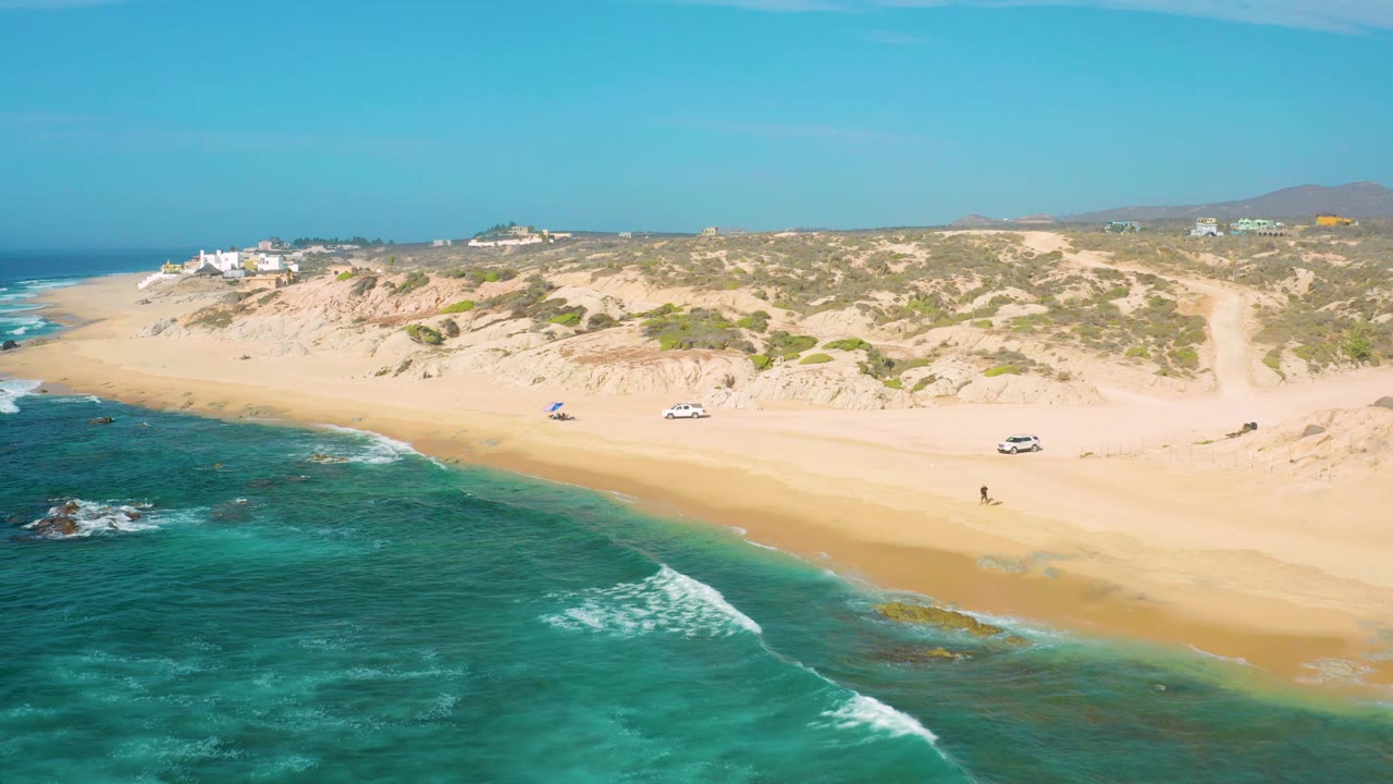 Shipwrecks Beach appears from an oblique drone angle with darker reef patches in the water, a clean sand band, scattered vehicles, small umbrellas, and homes or buildings above the dunes.