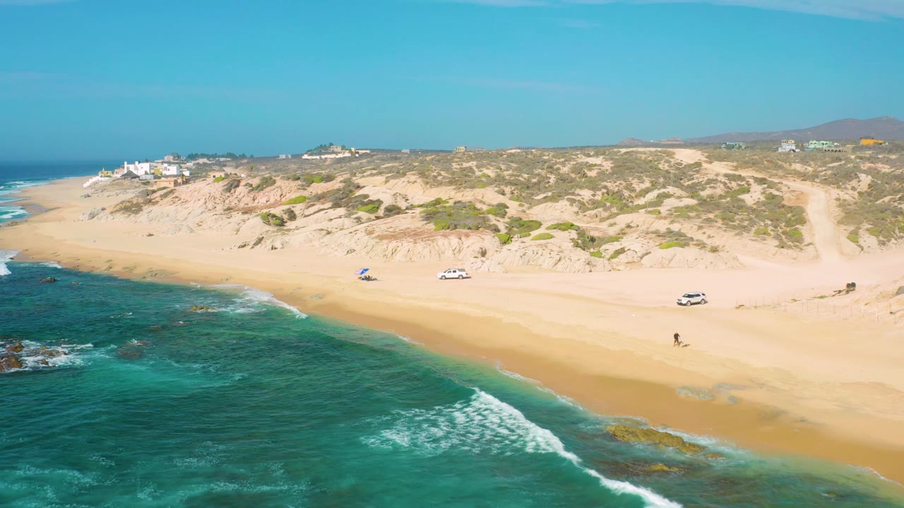 Aerial shoreline with green water, pale sand, one walker, cars, and dune scrub.