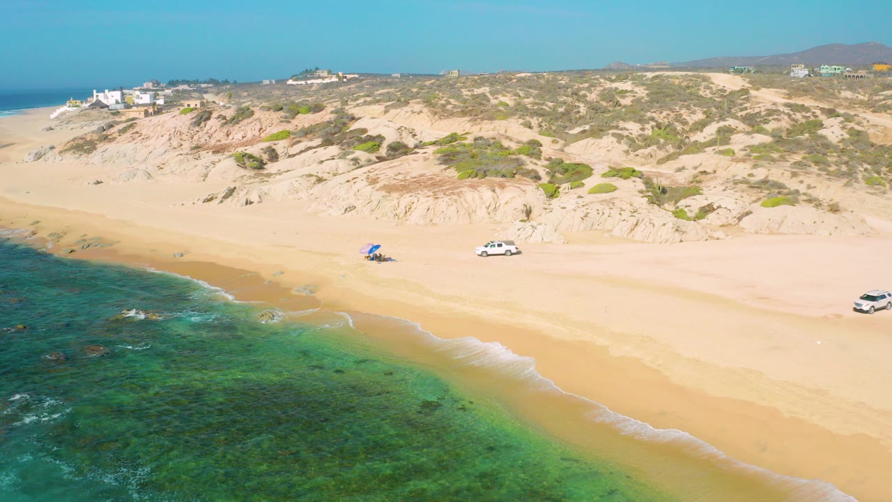 Aerial frame at Shipwrecks Beach, Baja California Sur, showing shallow green-blue water along tan sand, dunes behind, a white car, a blue beach shade, and a small visible person nearby.