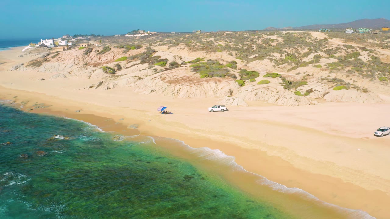 Wide beach aerial with clear water, low dunes, a white car, and a blue shade on sand.