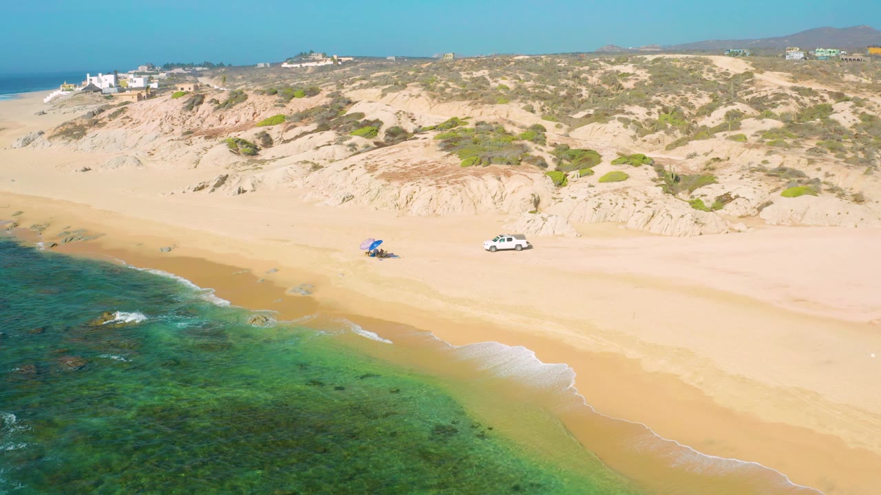At Shipwrecks Beach, rocky patches appear in the shallow green water while a white car and blue beach shade sit apart on the pale sand below the dune slope.