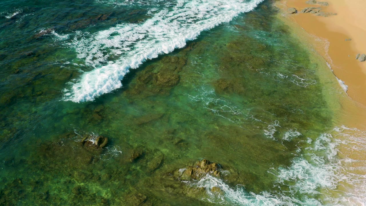 Overhead view at Shipwrecks Beach, Mexico, with white surf crossing green-blue shallows, rocks under the water, tan sand at right, and no visible people.