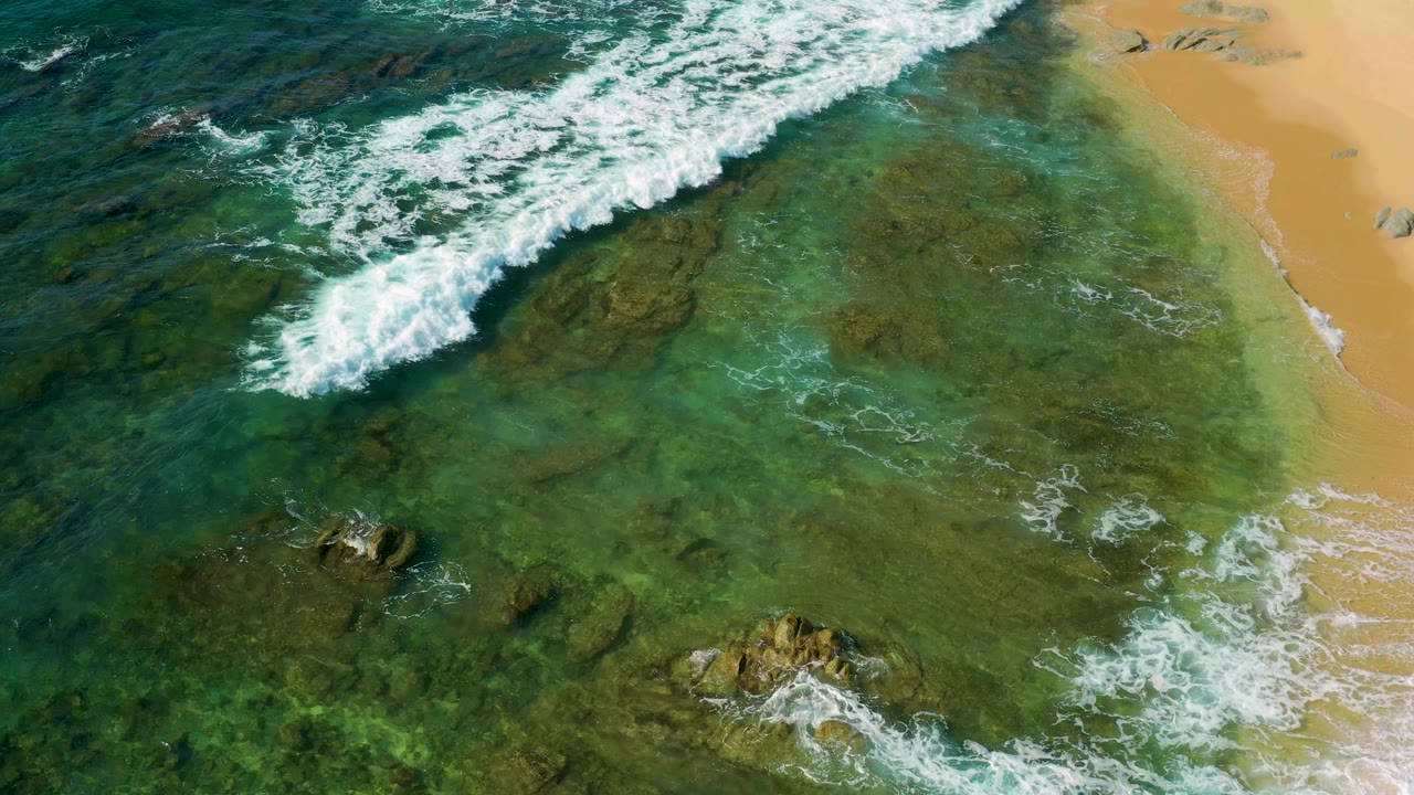 Aerial shoreline frame from Shipwrecks Beach, Mexico, with a broad white foam line over clear green water, submerged rocks below, sand at the right edge, and no visible people.