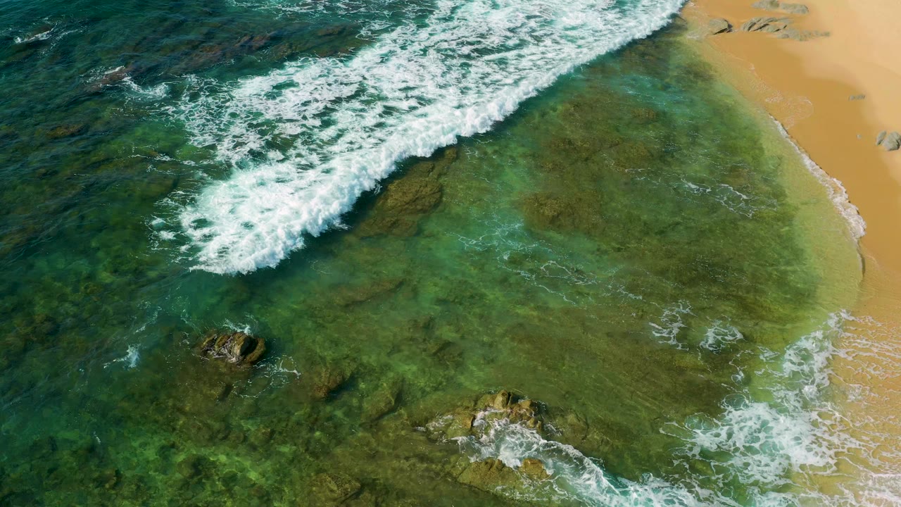 Top-down view at Shipwrecks Beach, Mexico, where diagonal surf foam advances across green shallow water, small rocks show below, sand borders the right side, and no visible people appear.