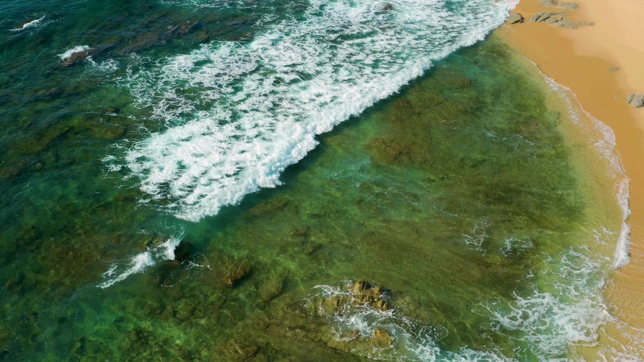 Overhead frame from Shipwrecks Beach, Mexico, with a larger white breaker crossing the upper half, clear green shallows below, rocks near the lower edge, sand at right, and no visible people.