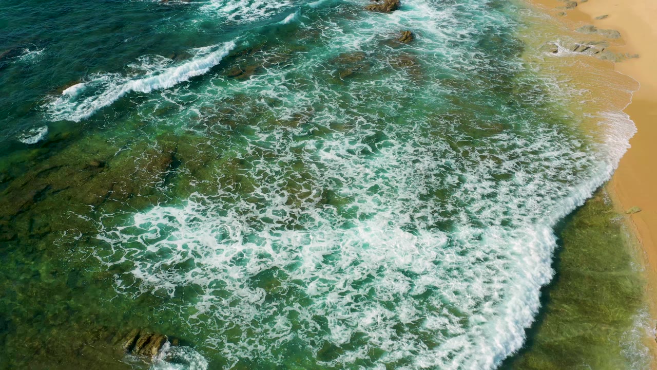 Aerial frame at Shipwrecks Beach, Baja California Sur, where broken wave foam crosses clear shallow water toward the sand, with dark rocks visible below and no visible people.