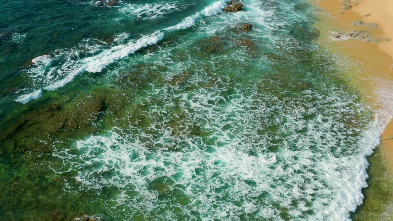Overhead view of Shipwrecks Beach, Baja California Sur, with a curved white surf band, exposed rocks near the upper water, amber sand on the right, and no visible people.