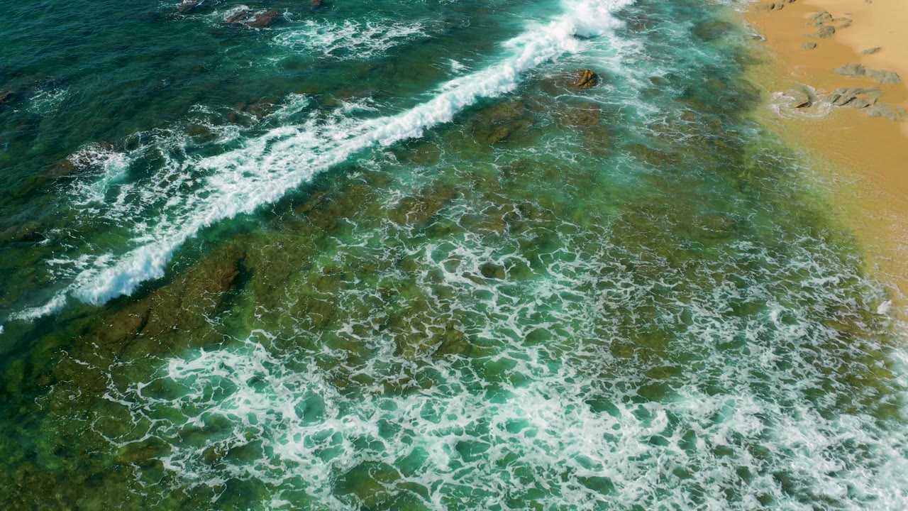 Aerial view over Shipwrecks Beach, Baja California Sur, with a diagonal wave breaking from upper center toward the lower edge, rocky patches beneath green water, sand at right, and no visible people.