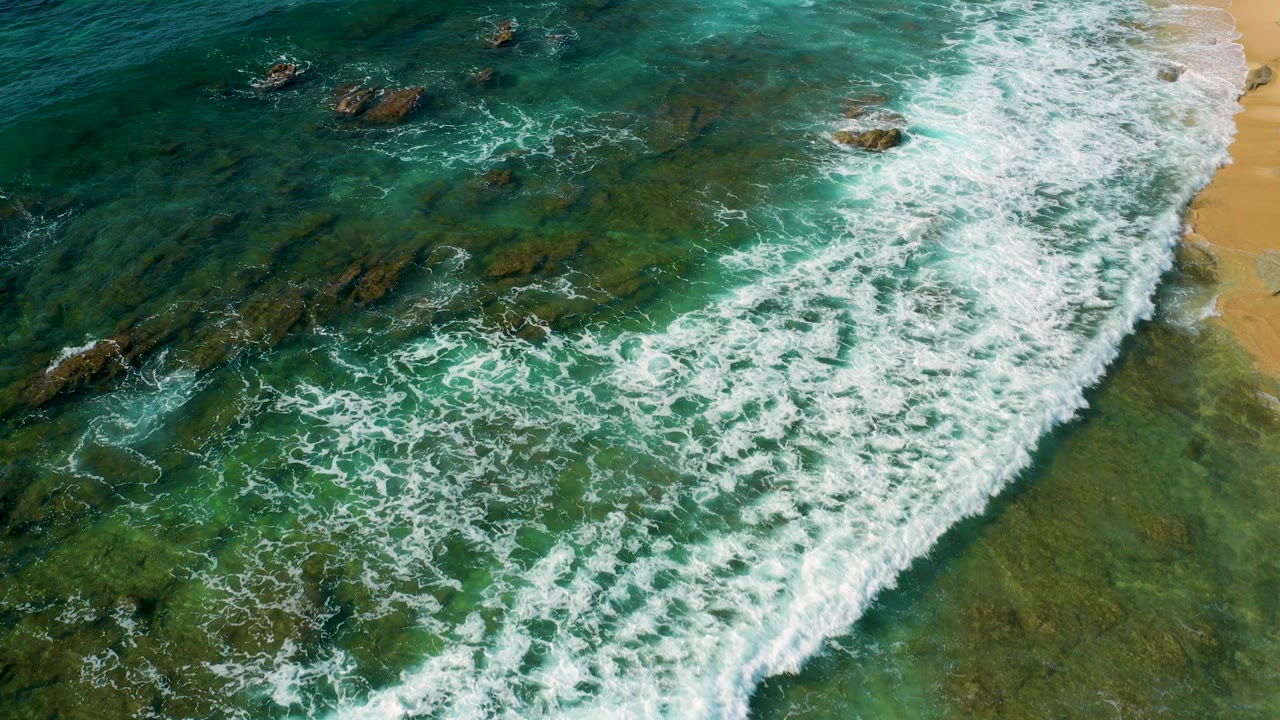 Aerial view at Shipwrecks Beach in Baja California Sur with white surf spreading over green-blue water, dark rocks below the surface, a sandy edge at right, and no visible people.