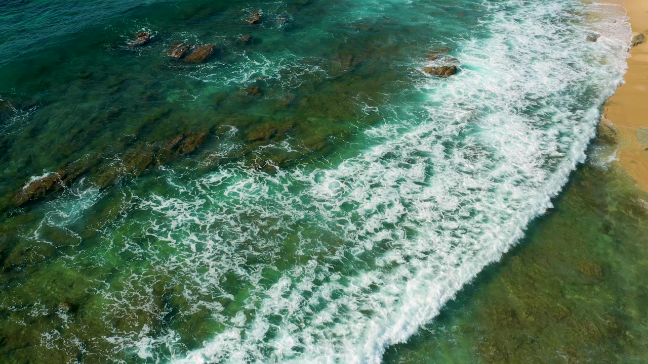 Aerial frame from Shipwrecks Beach with a broad white wave band crossing shallow turquoise water, dark rock shelves visible beneath the surface, and no visible people.