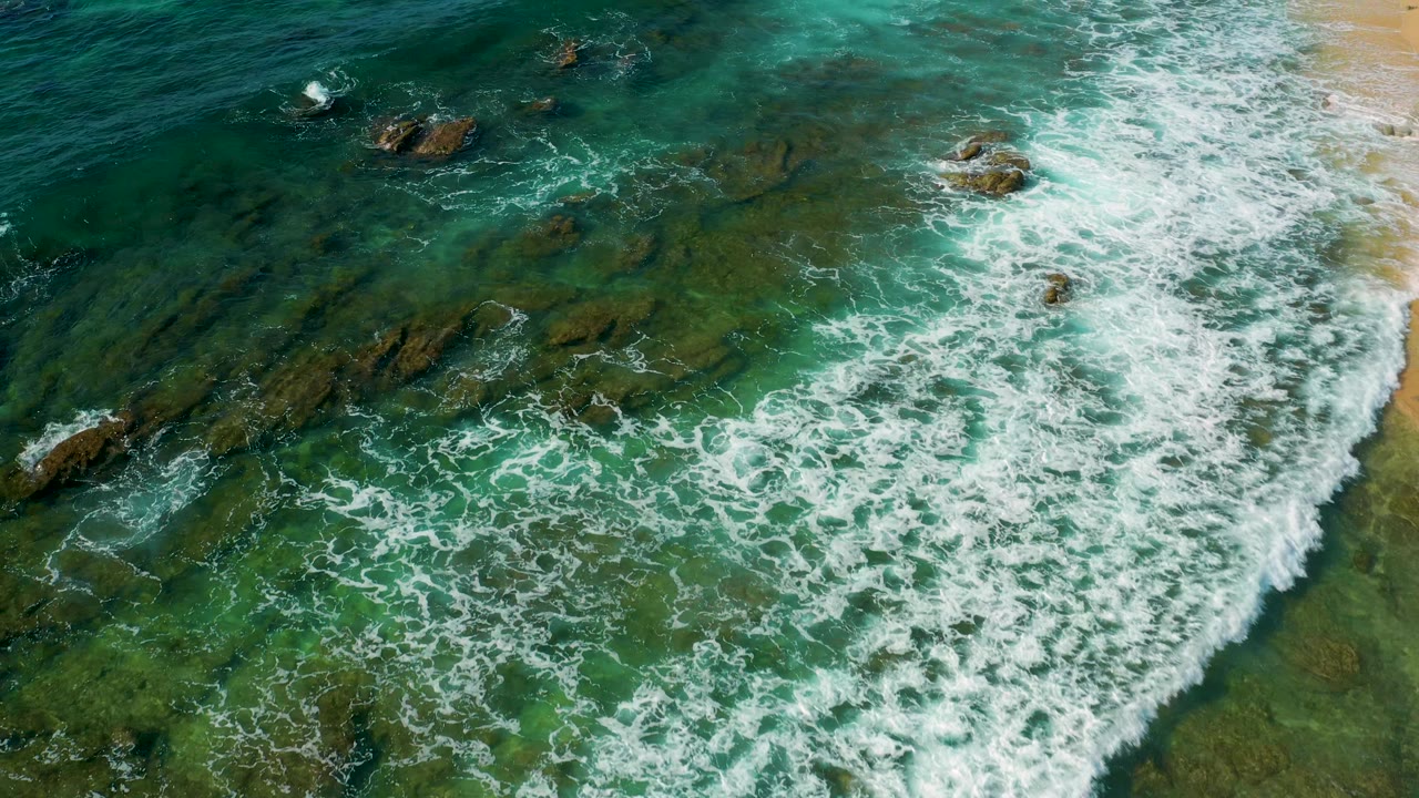 Aerial view at Shipwrecks Beach showing foamy surf breaking across scattered rocks, clear green-blue water, sand along the upper right edge, and no visible people.