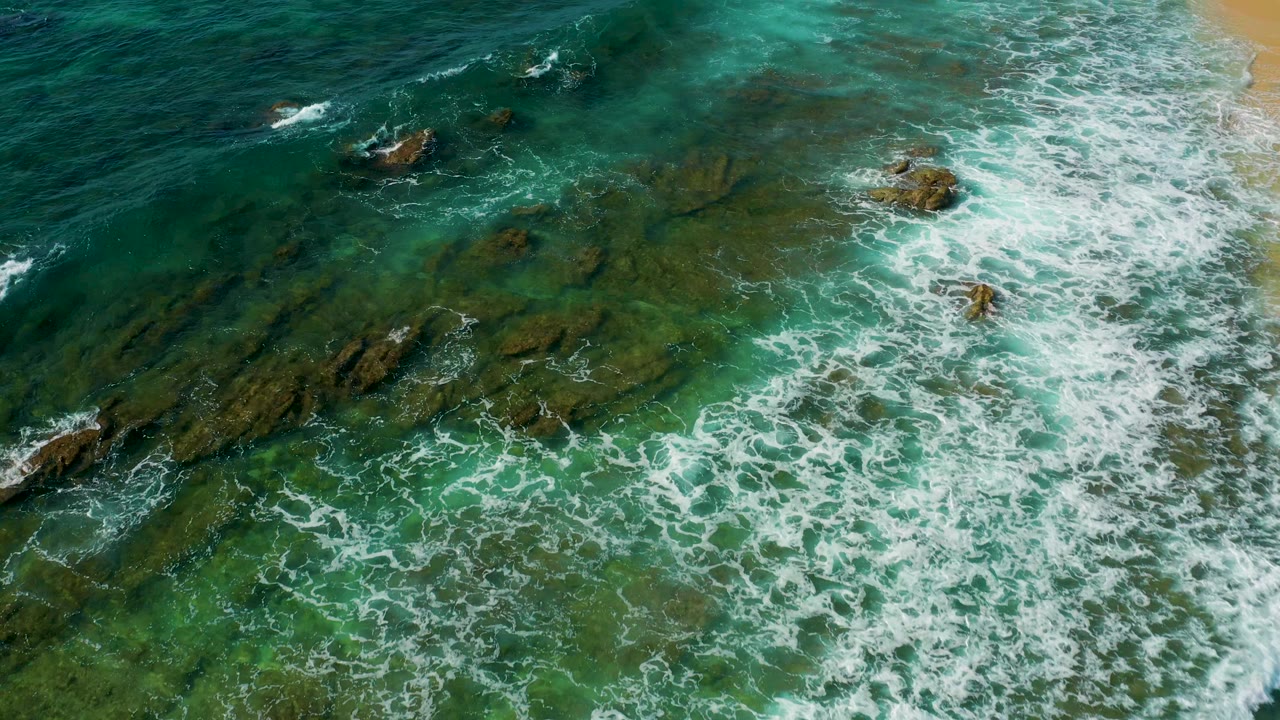 Shipwrecks Beach aerial frame with dense whitewater filling the lower half, green shallows beneath, exposed rocks near the beach edge, and no visible people.