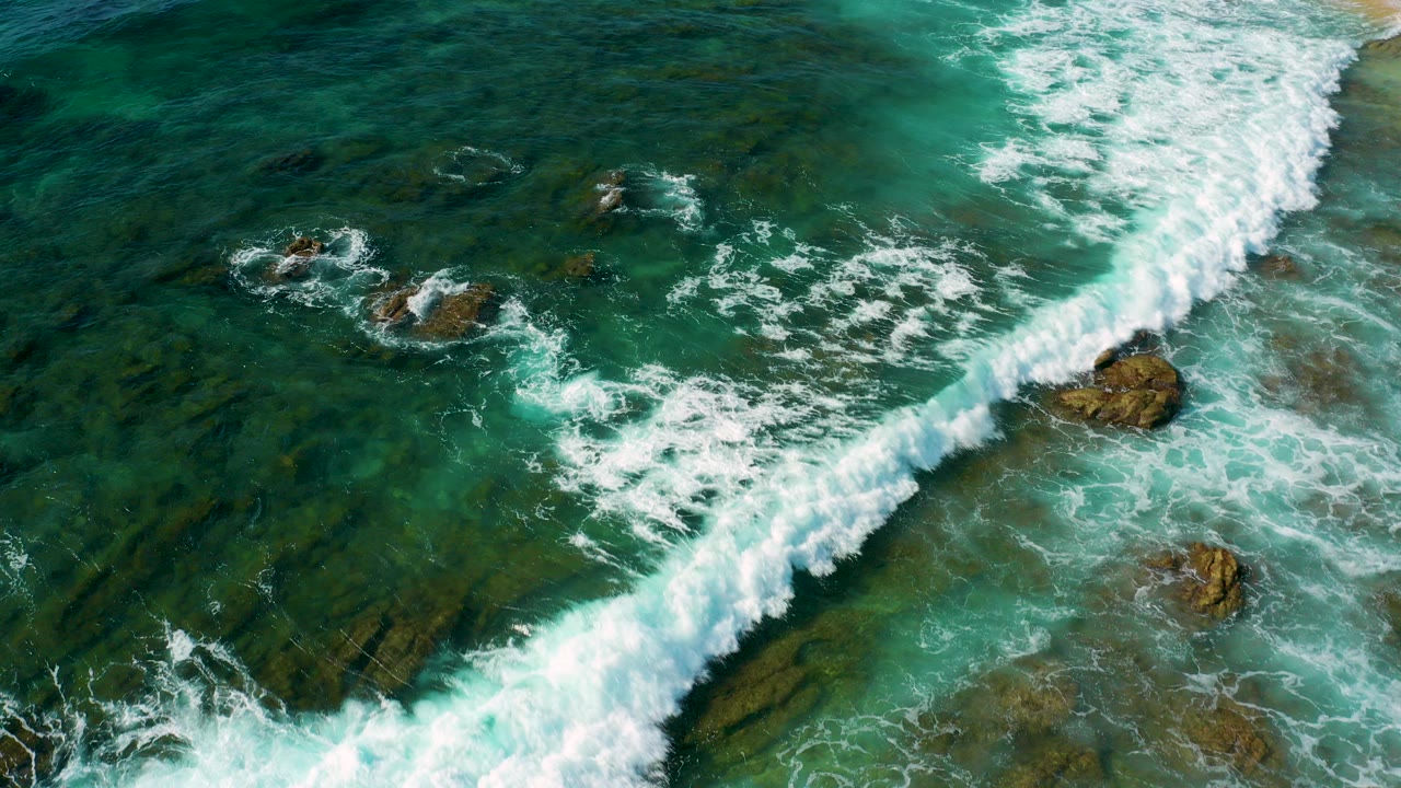 Aerial view at Shipwrecks Beach, Baja California Sur, showing a white surf line crossing blue-green water and jagged rocks, with no visible people.