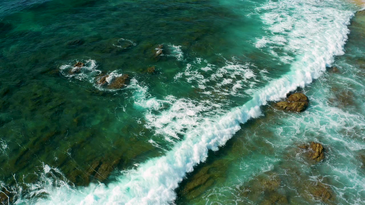 Aerial frame at Shipwrecks Beach with a diagonal band of whitewater moving over shallow rock shelves and scattered exposed rocks, with no visible people.