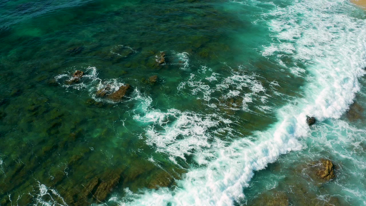 Overhead view of Shipwrecks Beach surf curling around brown rocks in blue-green water, with broken white foam and no visible people.