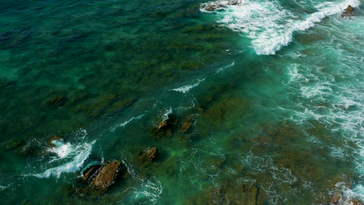Aerial view at Shipwrecks Beach, Baja California Sur, showing green shallow water, submerged rocky bottom, scattered exposed rocks, and white surf foam with no visible people.