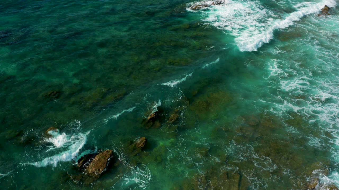 White wave wash crossing green shallow water around dark rocks.