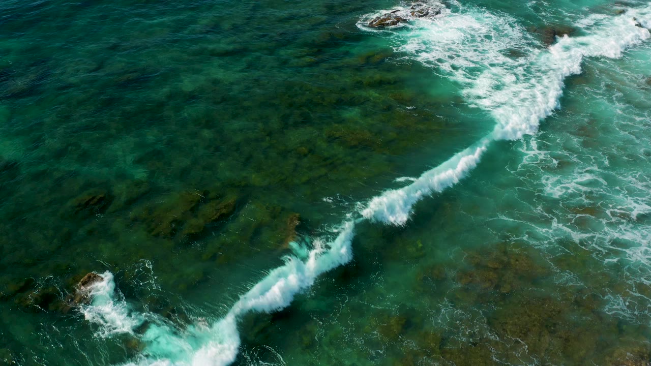 Aerial view at Shipwrecks Beach where a diagonal line of white breaking surf passes over clear green water and algae-darkened rock shelves, with no visible people.
