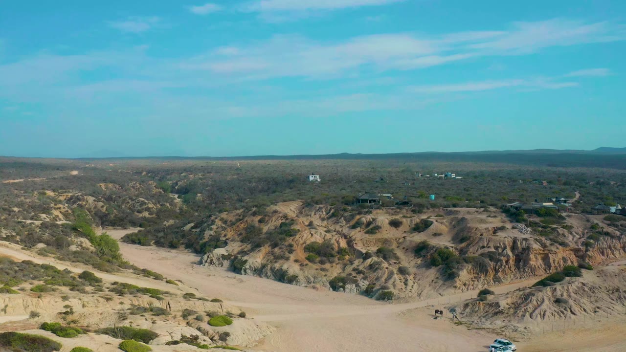 Elevated desert landscape with sandy foreground, scrub ridges, dirt tracks, and distant buildings.