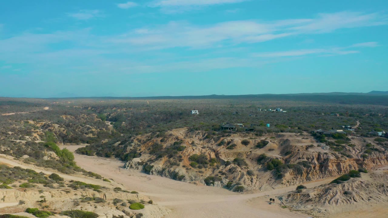 At 0:02, the aerial view shows Shipwrecks Beach terrain with tan sand, scattered green bushes, small distant structures, a level horizon, and no visible people.